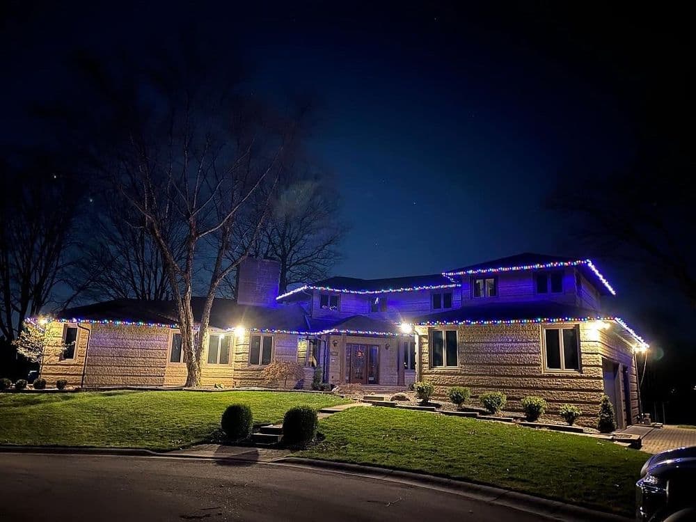 Modern home at night adorned with blue and white holiday lights, surrounded by landscaped lawn.