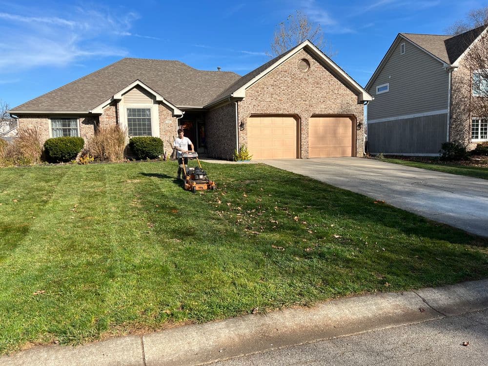 Person mowing a lawn in front of a suburban home with a brick facade and double garage.